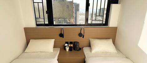 Una habitación de hotel compacta con dos camas individuales, una mesita con cafetera y un ventanal con vistas a la ciudad.