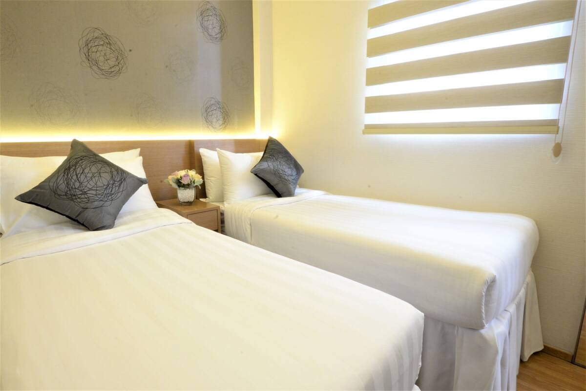 Ein Hotelzimmer mit zwei Betten, beide mit weißen Laken und grauen, gemusterten Kissen. Dazwischen befindet sich ein Nachttisch mit einer Blumenvase und einer Lampe.