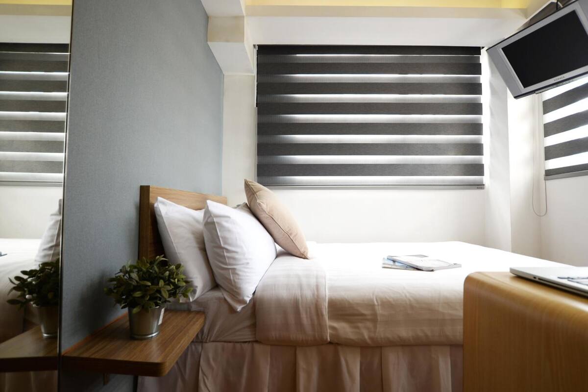 Ein Hotelzimmer mit einem Bett, einem Nachttisch mit Pflanze, einem Fernseher und einem Fenster mit Jalousien.