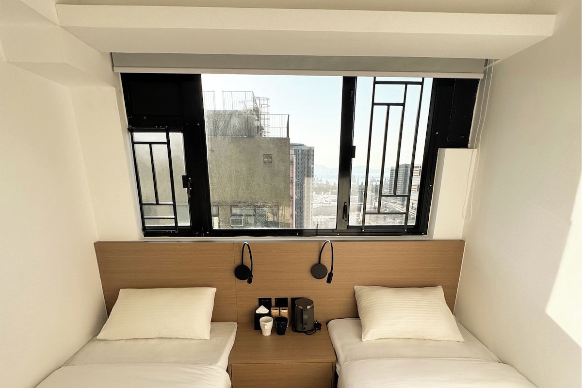 A hotel room with two beds, a window with a view of buildings, a wooden headboard, and a small table with a coffee maker.