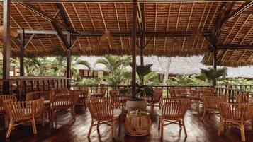 Un espacio al aire libre con sillas de mimbre y techo de paja, con vista a un jardín con palmeras.