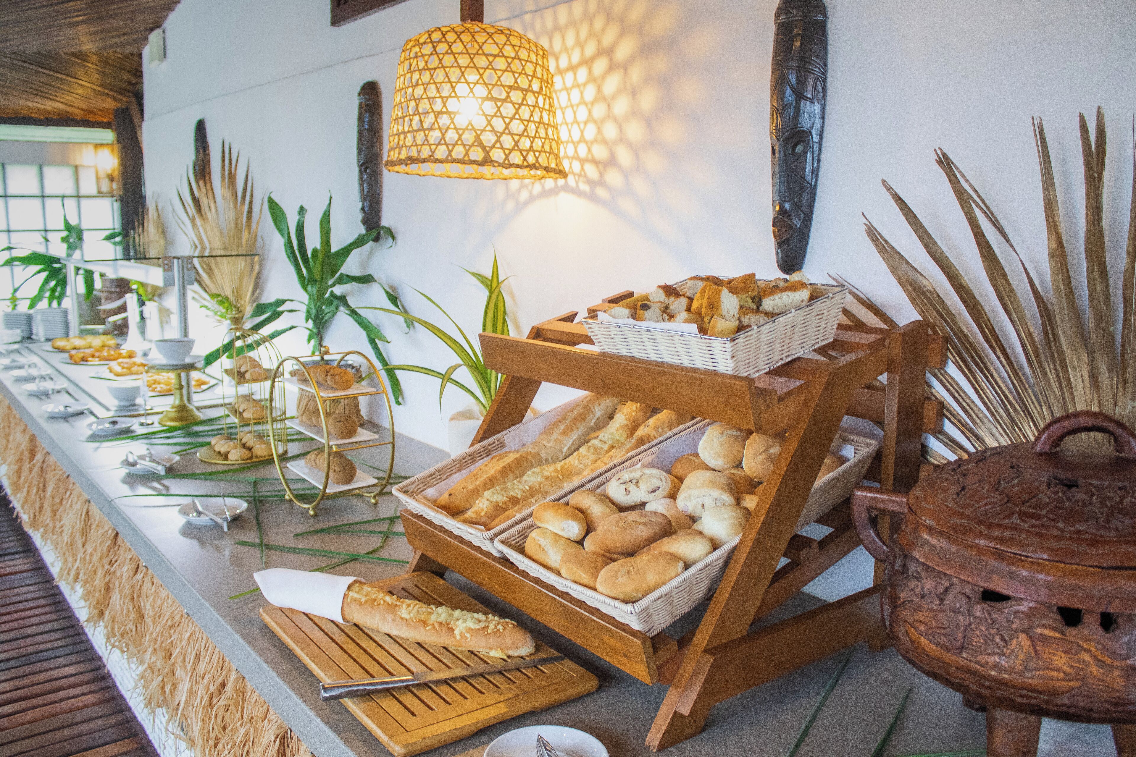Una mesa de buffet con panes y pastelería variada, un soporte de madera y plantas decorativas.