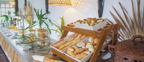 Una mesa de buffet con panes y pastelería variada, un soporte de madera y plantas decorativas.