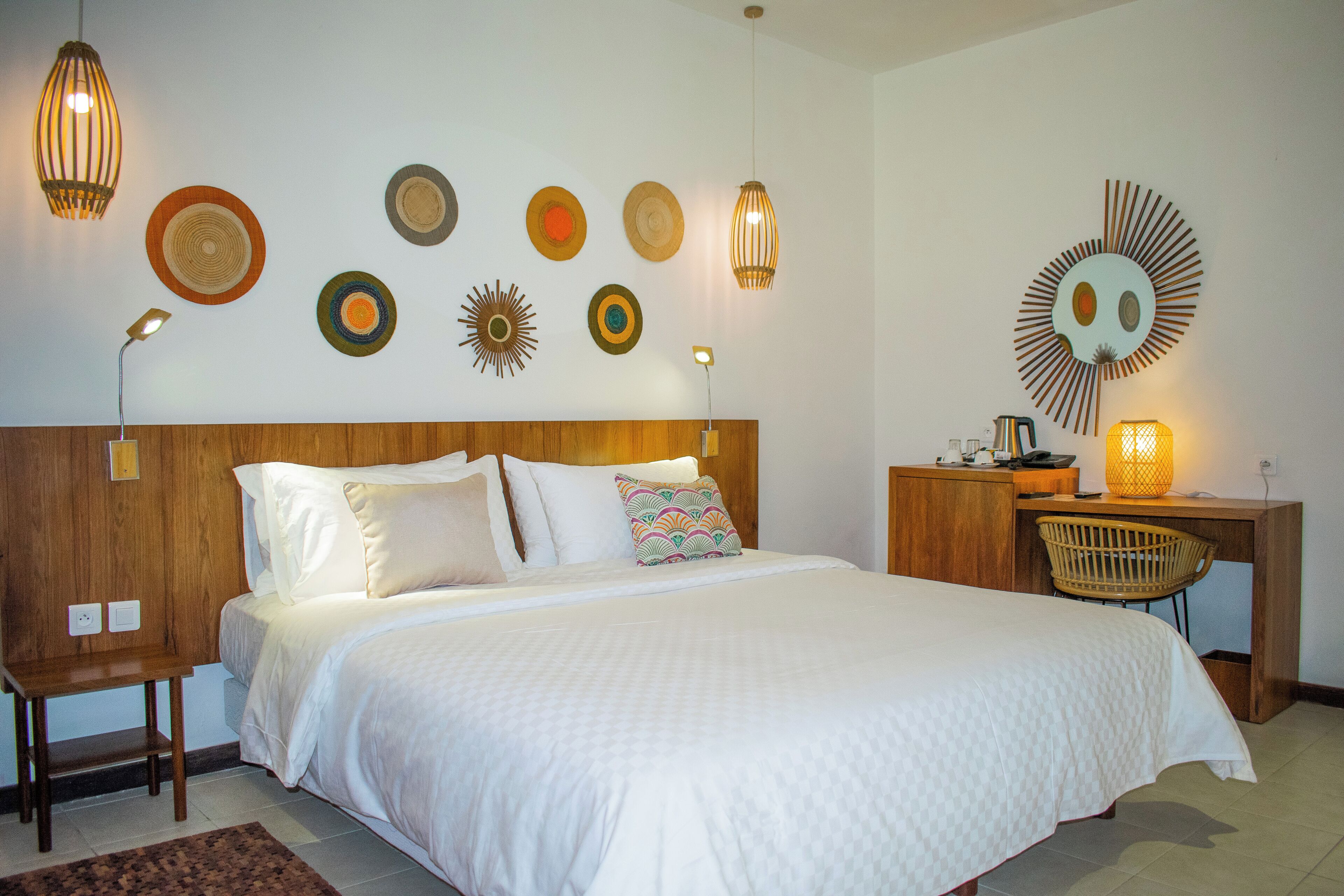 A bedroom with a wooden headboard, a bed with white bedding, a desk with a chair, and wall decor including plates and a sun-like design.