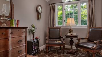 A room with a wooden dresser, two leather chairs, a table with a lamp, a clock, and a window with curtains.