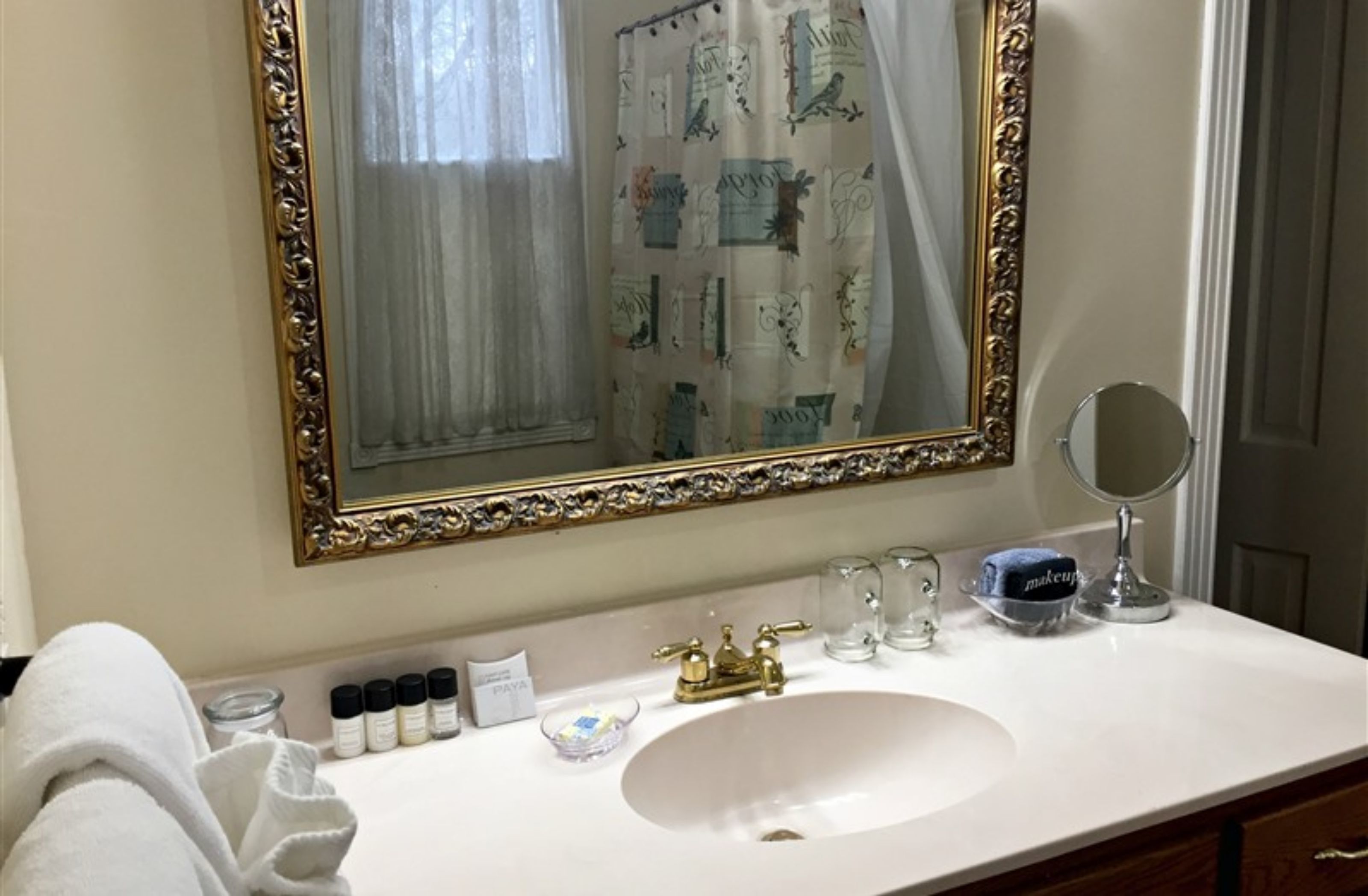 A bathroom with a white sink, gold faucets, a large mirror with an ornate frame, and a shower curtain with a floral pattern.
