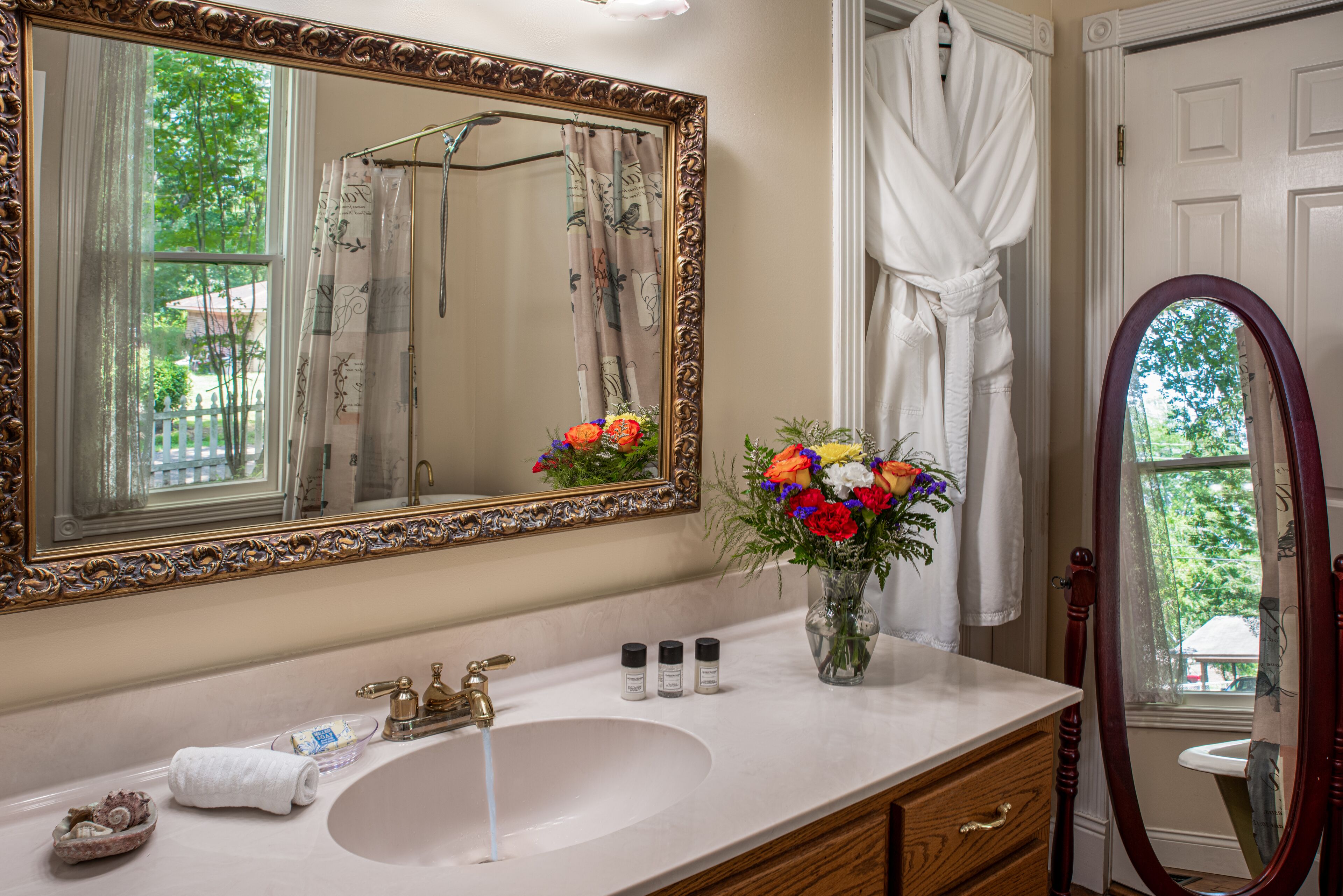 Une salle de bain avec un grand miroir, un lavabo, un vase de fleurs et une vue sur l’extérieur.