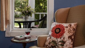 Una habitación acogedora con vistas a un jardín a través de una ventana, una pequeña mesa redonda con un libro y una copa de vino tinto, y un sofá con un cojín de girasol.
