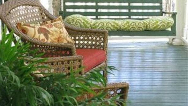 Una silla de mimbre con un cojín rojo y una almohada estampada, ubicada frente a un banco verde con un cojín a juego.