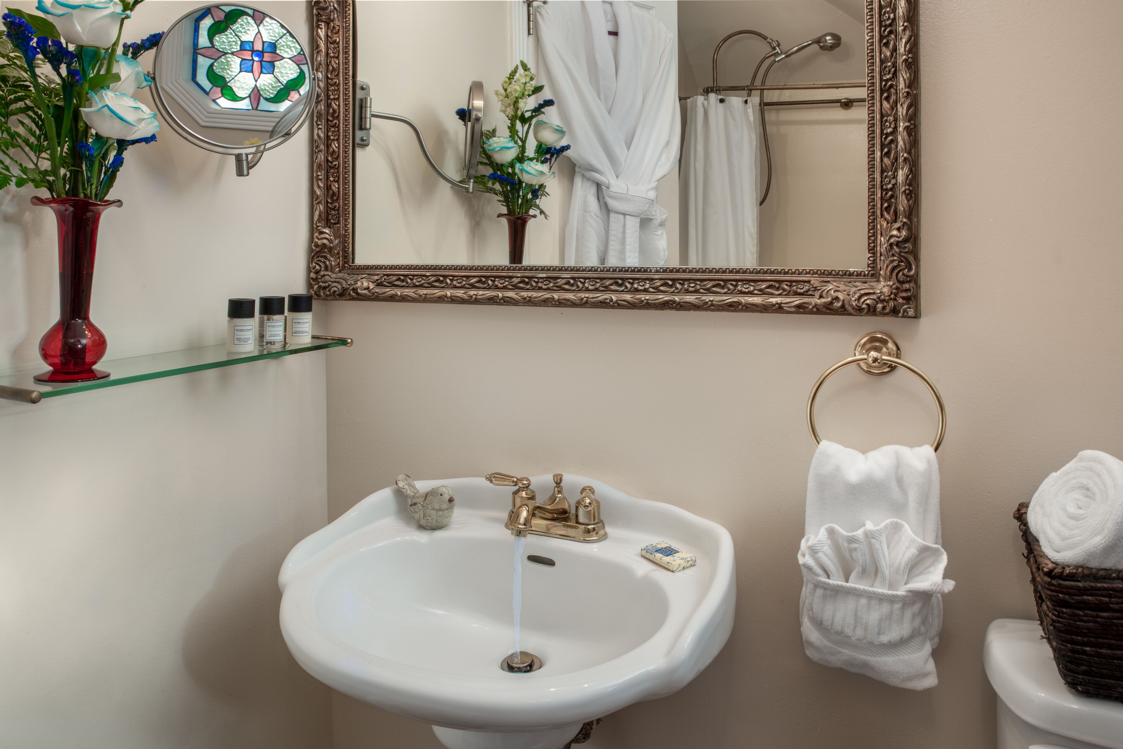 Un baño con un lavamanos blanco, un espejo, un florero con flores y un perchero para toallas.