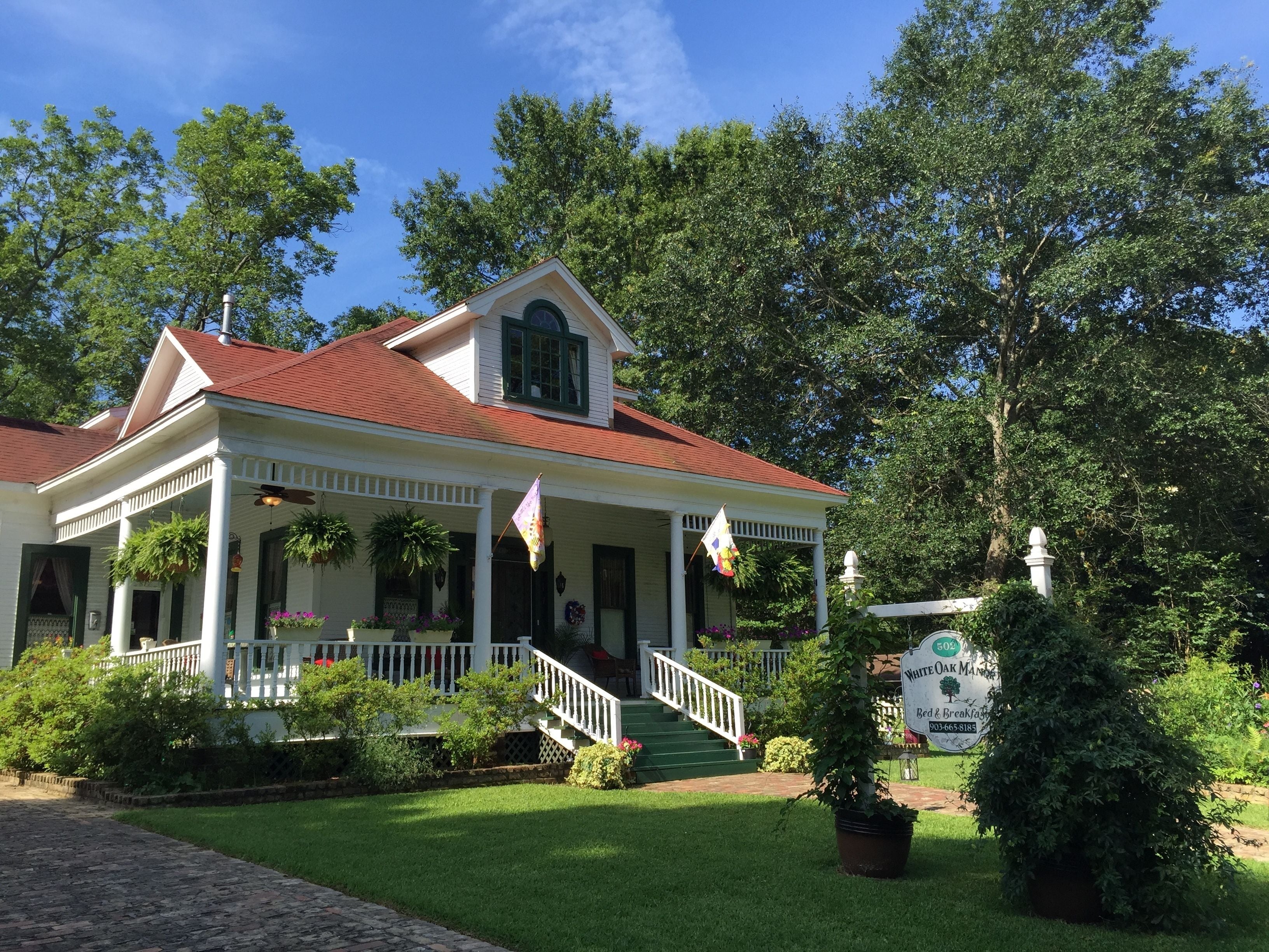 A white colonial-style building with a red roof, a porch with hanging plants, and a sign indicating it is a bed and breakfast.