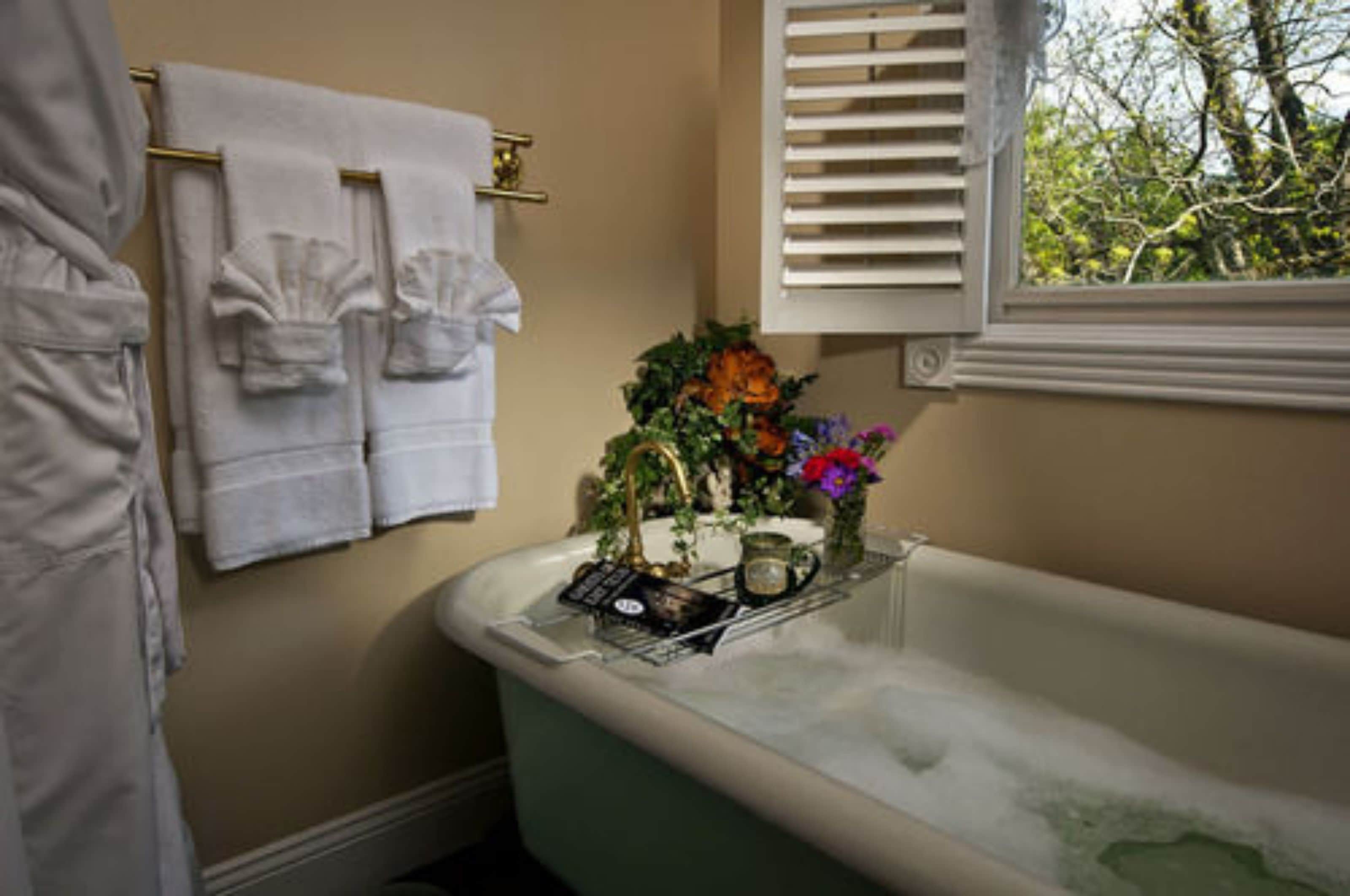 A bathroom with a bathtub, towels, a window with blinds, and a vase with flowers.