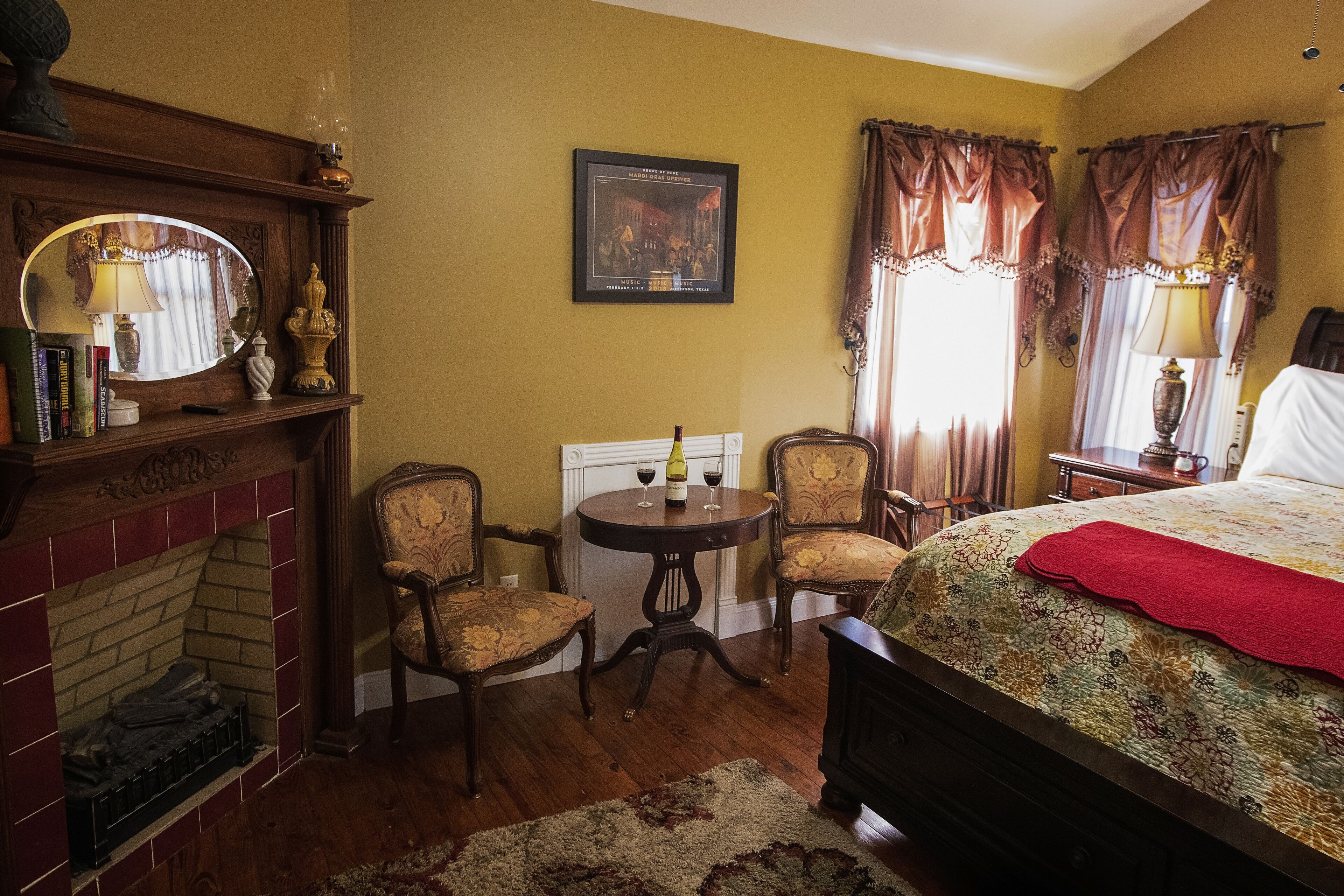 A room with a fireplace, two armchairs, a bed with a red blanket, a round table with a bottle and wine glasses, and a framed picture on the wall.
