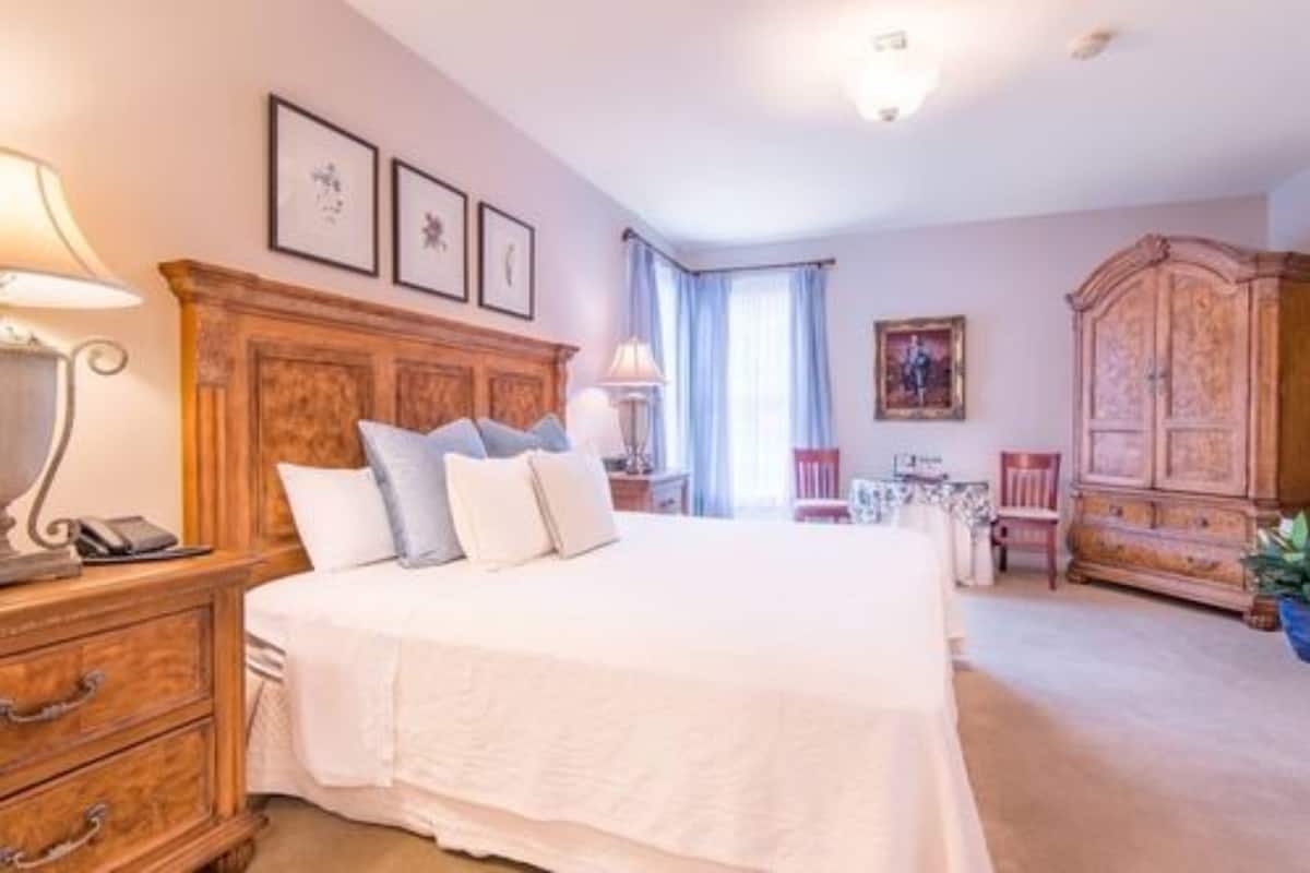 A bedroom with a wooden headboard, a bed with white bedding, a nightstand, a lamp, a dresser, a chair, a cabinet, and a window with curtains.