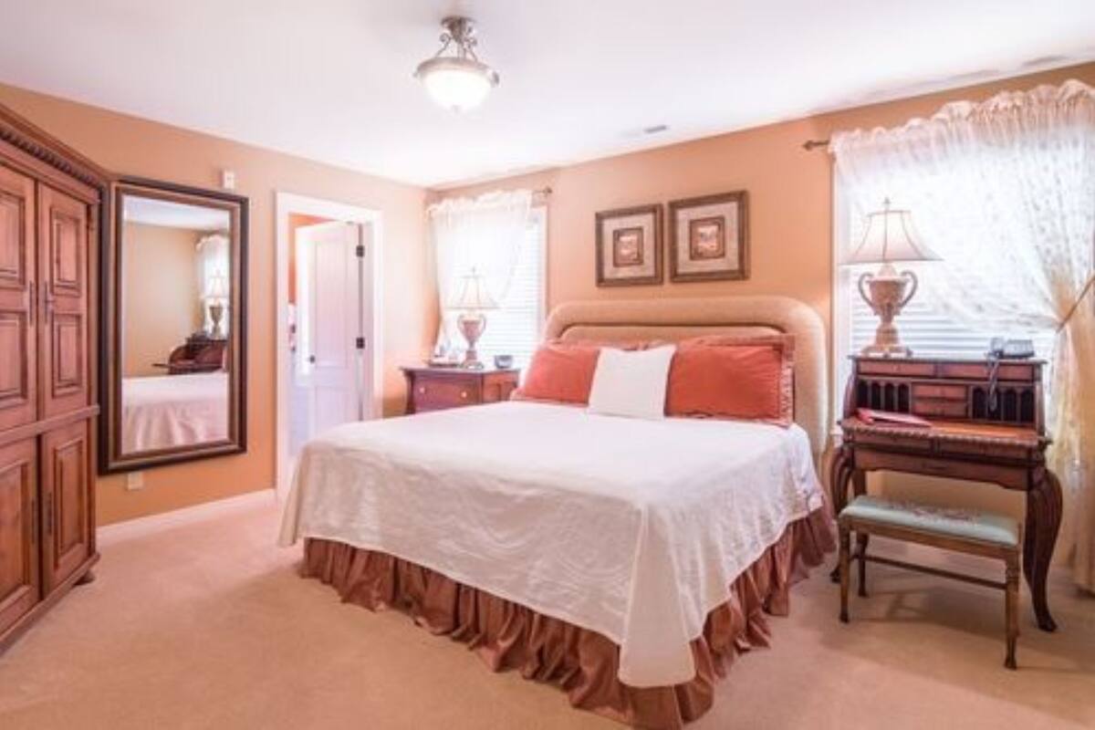 A bedroom with a bed, a wooden nightstand, a chair, a mirror, and a window with curtains.