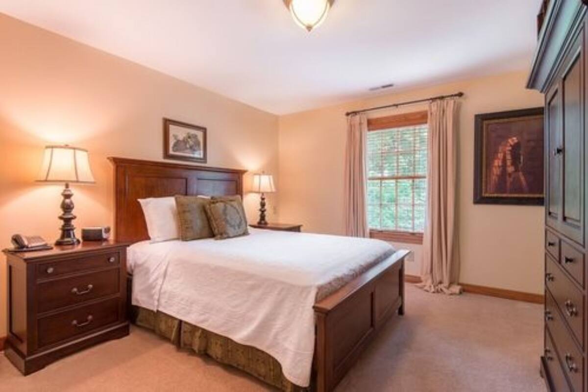 A bedroom with a large bed, two bedside tables with lamps, a dresser, and a window with curtains.