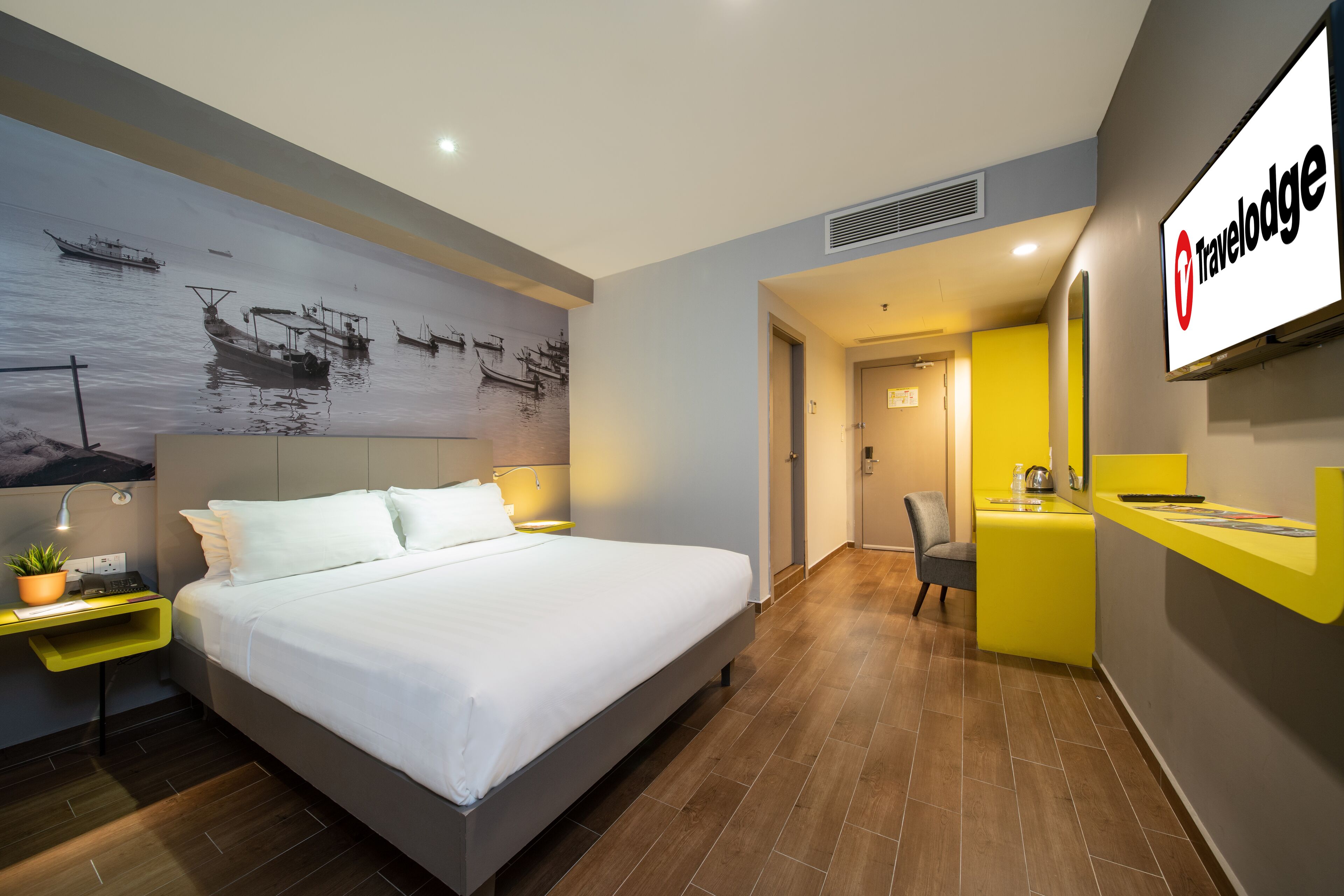 Ein modernes Hotelzimmer mit einem großen Bett, einem Schreibtisch mit Lampe, einem an der Wand befestigten Fernseher und einer Rezeption mit gelbem Empfangstresen.