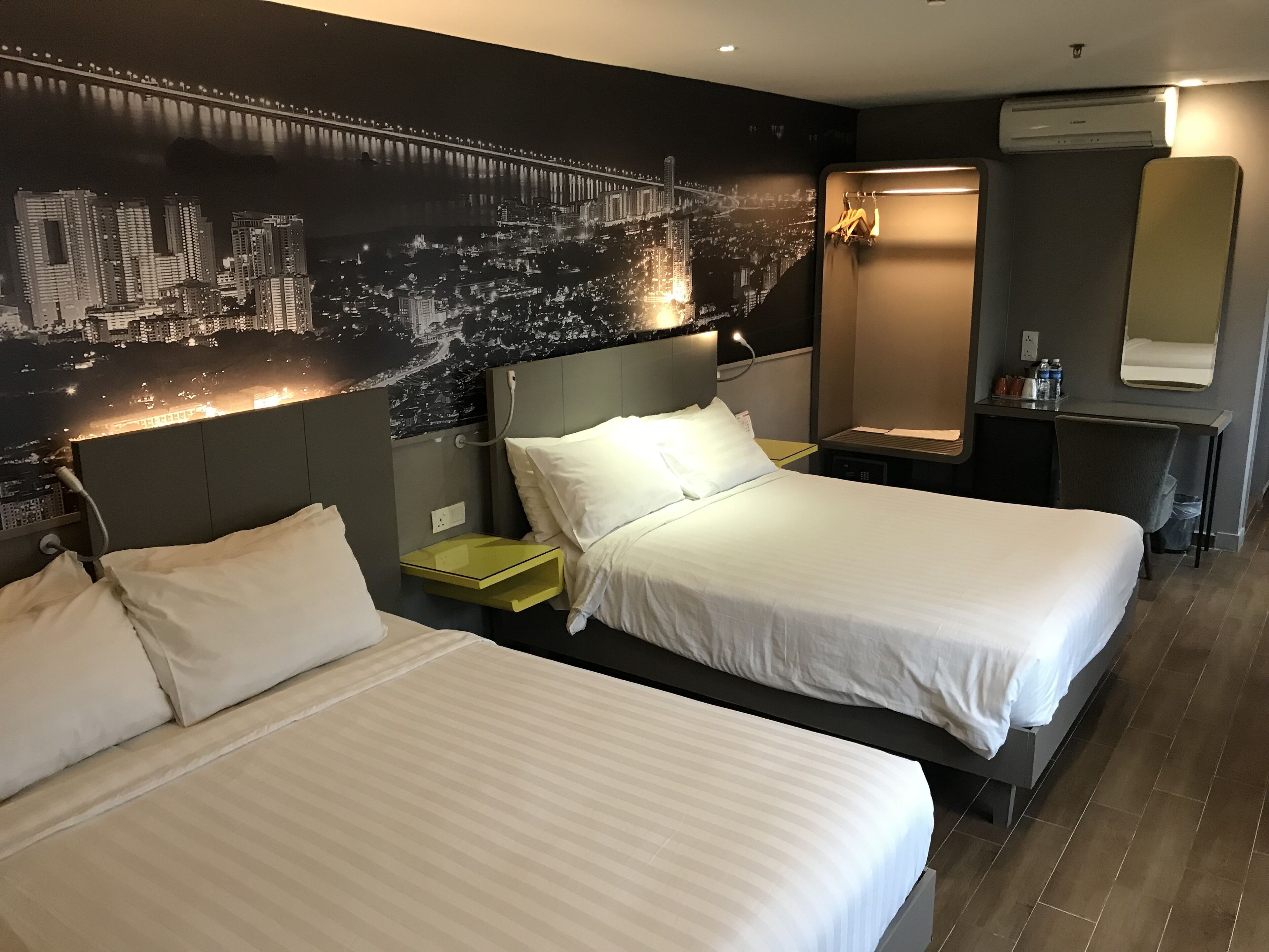 A hotel room with two beds, a nightstand, a desk, a chair, a mirror, and a wall mural of a cityscape.