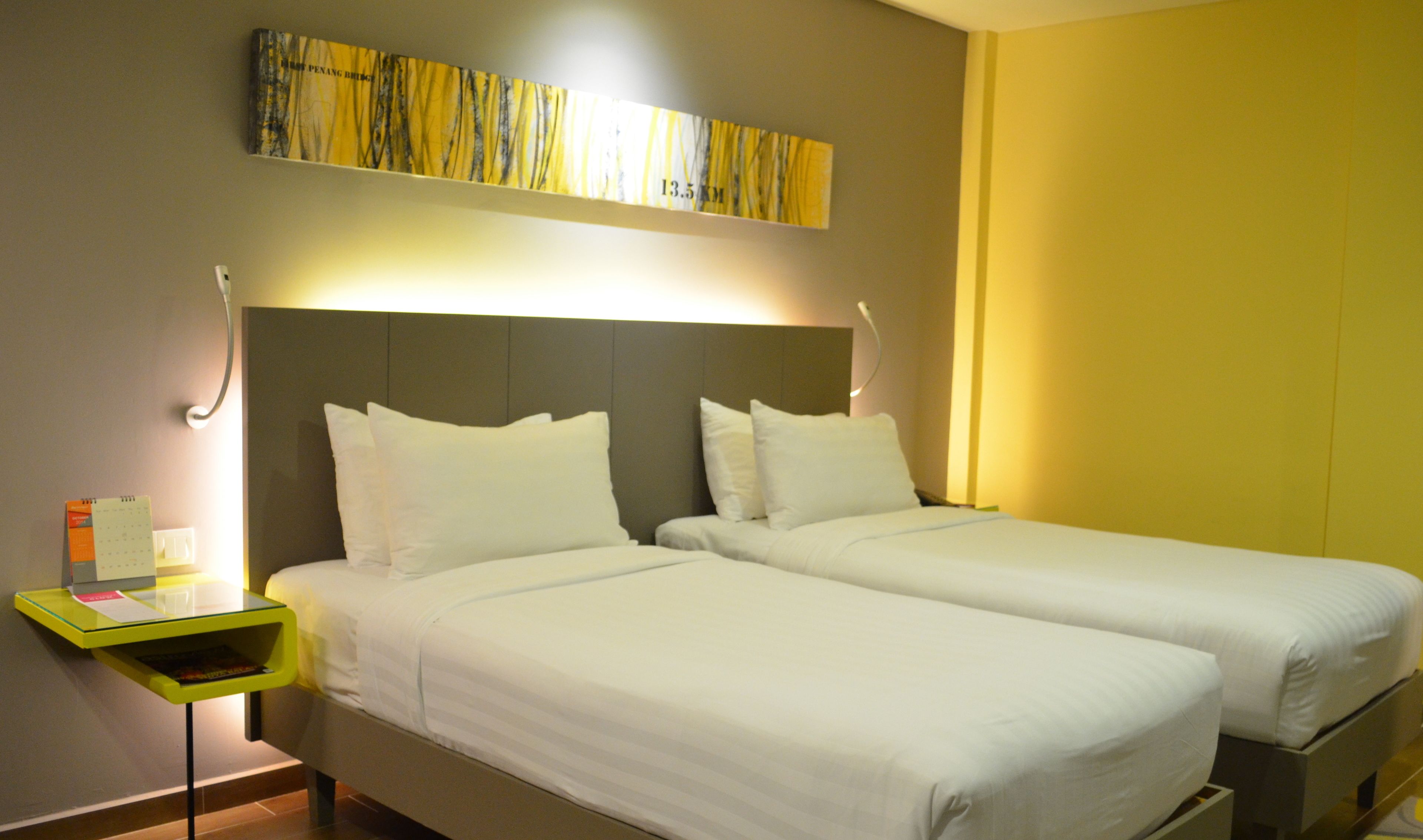 A hotel room with two single beds, a small bedside table with a lamp, a wall-mounted artwork, and a yellow accent wall.