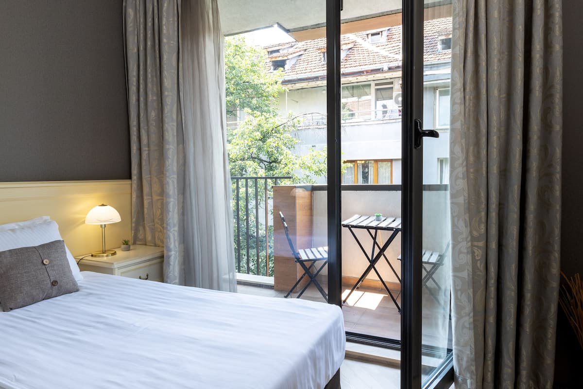 A hotel room with a bed, a nightstand with a lamp, a balcony with a table and chairs, and a view of the outside buildings and trees.