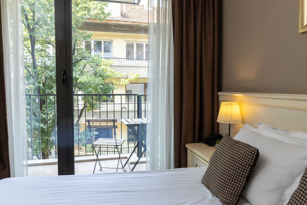 A hotel room with a bed, a nightstand, a lamp, and a view of a balcony with a table and chairs.