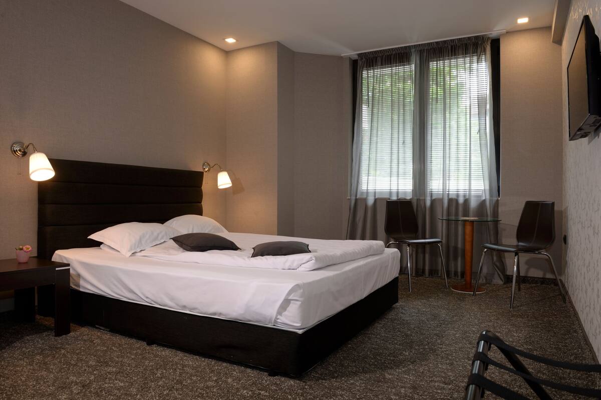 A hotel room with a large bed, two bedside tables, a small table with chairs, and a window with curtains.