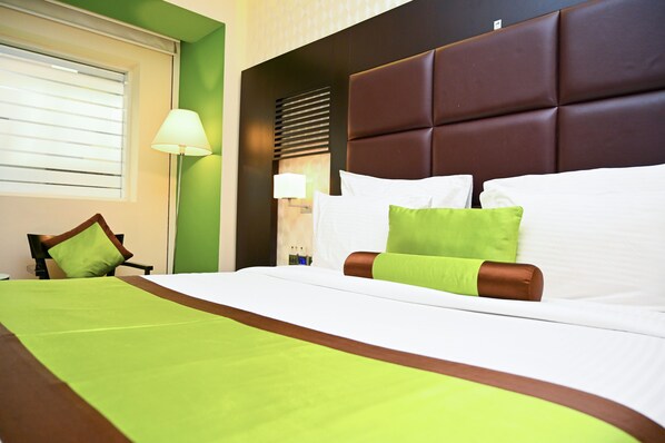A hotel room with a large bed, green and brown bedding, a headboard, a window with blinds, and a lamp.
