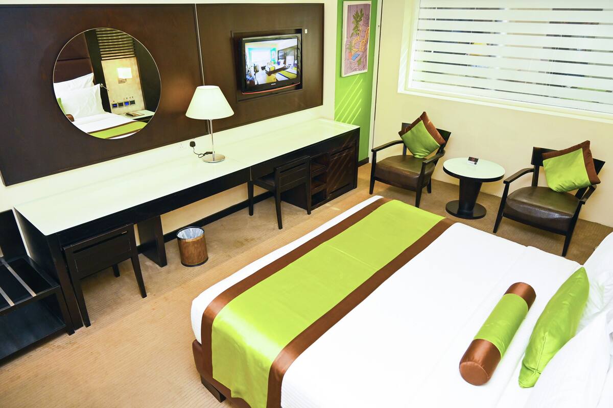 A hotel room with a bed, a desk, a chair, a small table, a TV, and a window with blinds.