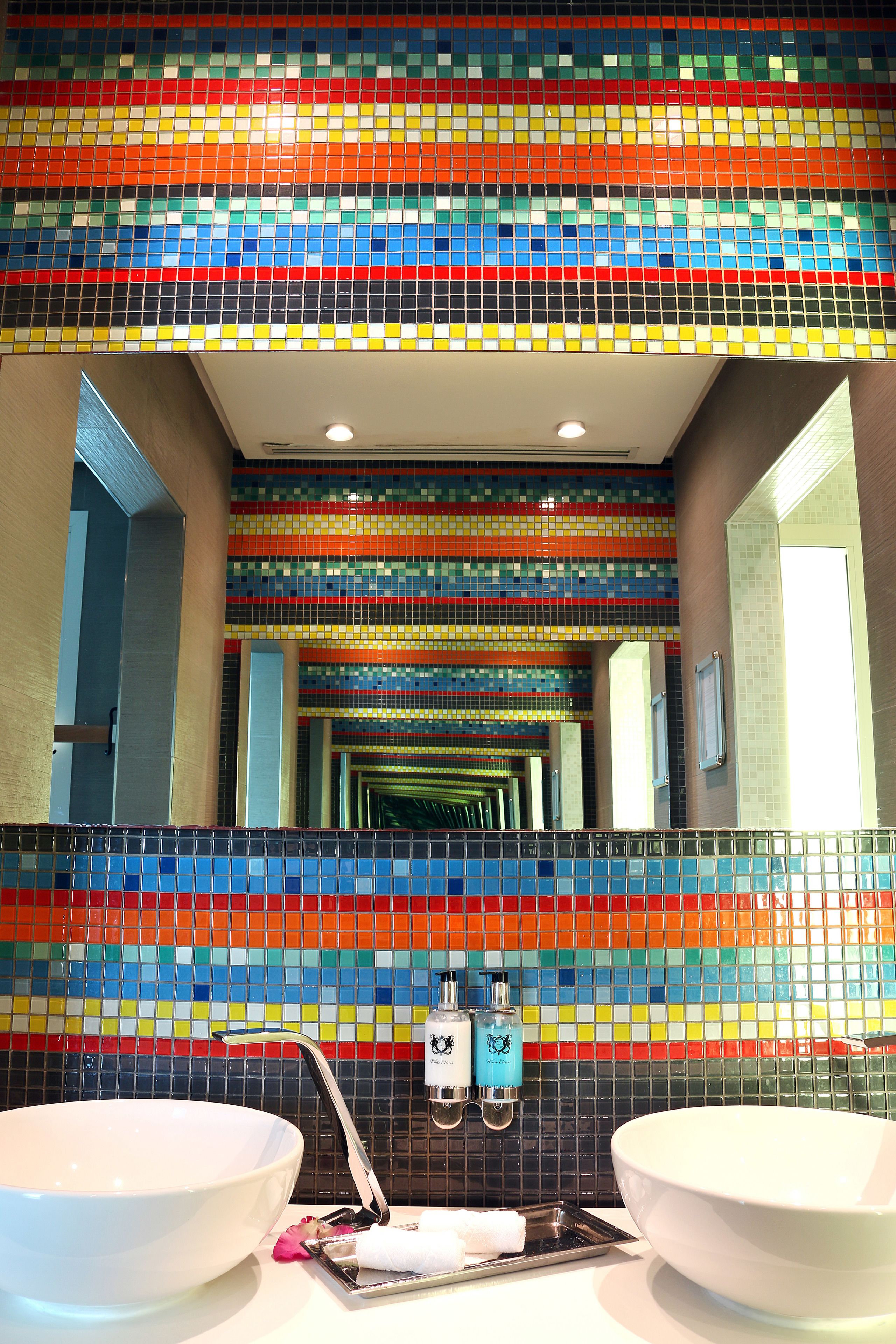 A bathroom with two sinks, colorful mosaic tiles on the walls, and a mirror reflecting the space.