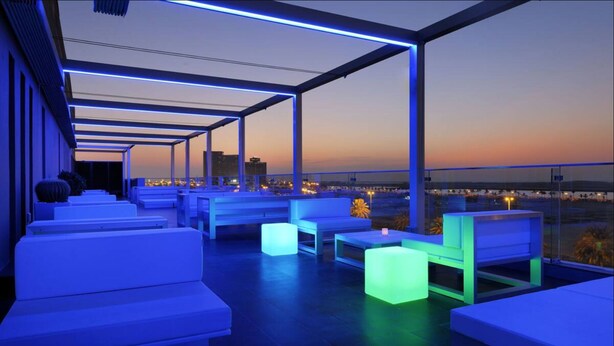 Un lounge en la azotea con asientos blancos y mesas iluminadas, con vistas a la ciudad al atardecer.