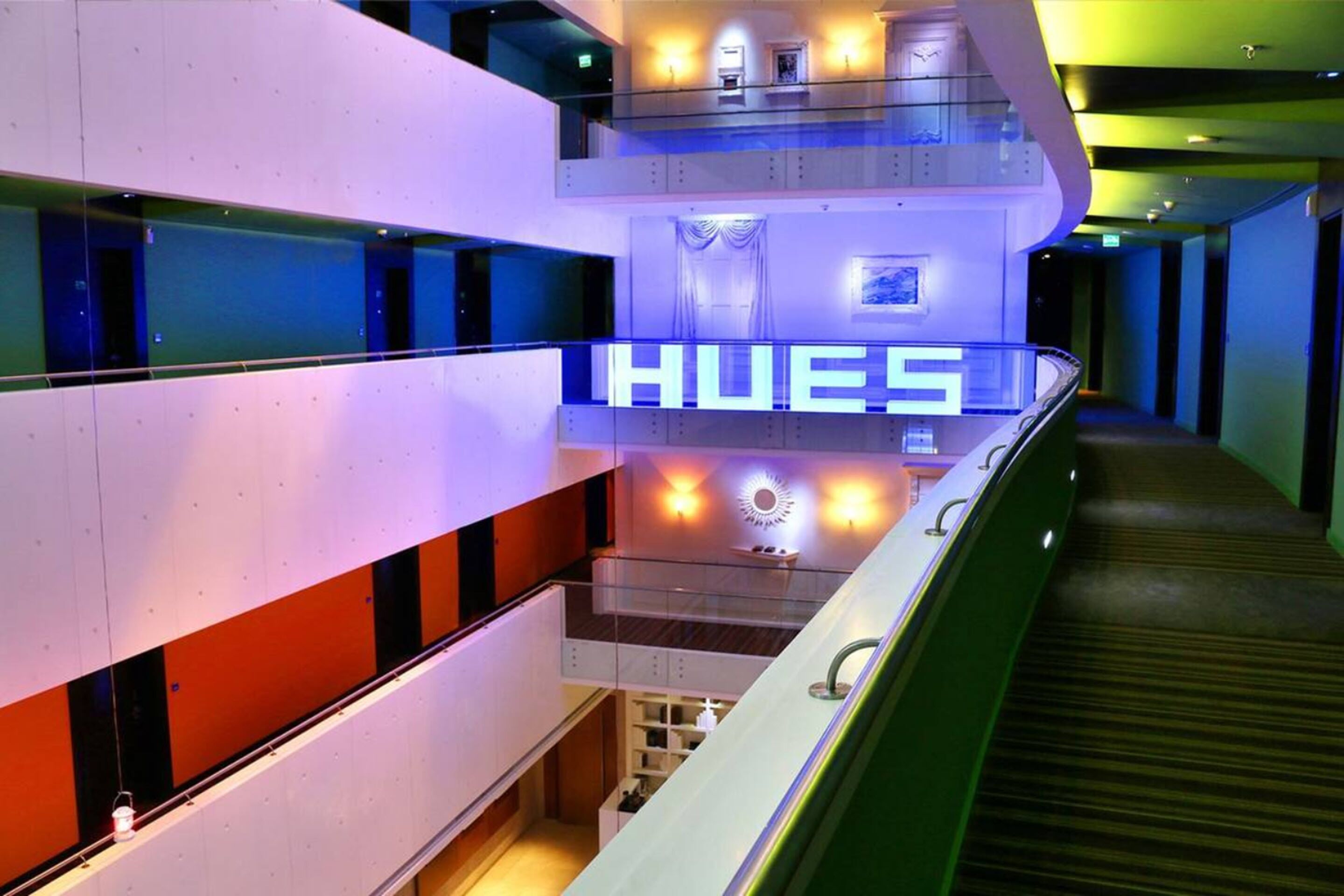 Un intérieur d’hôtel moderne, avec une enseigne bien visible portant le nom « HUES », de nombreux étages et un escalier.