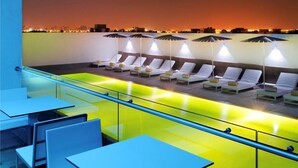 Un área de lounge en la azotea con sillones de sol y sombrillas, una piscina con borde amarillo y vistas a la ciudad al atardecer.