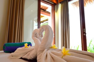 Uma cama bem arrumada com uma toalha dobrada em forma de cisne, uma toalha verde e roxa, e flores amarelas sobre a cama.