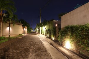 Rua pavimentada, ladeada por edifícios e áreas verdes, iluminada por postes de luz.