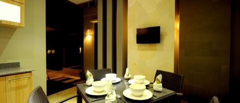 A dining area with a table set for four, surrounded by wicker chairs, a flat-screen TV mounted on the wall, and a kitchen area visible in the background.