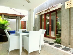 An outdoor patio with white wicker furniture, a table, and chairs. There is a potted plant and a wooden door in the background. - Jas Boutique Villas (Seminyak)
