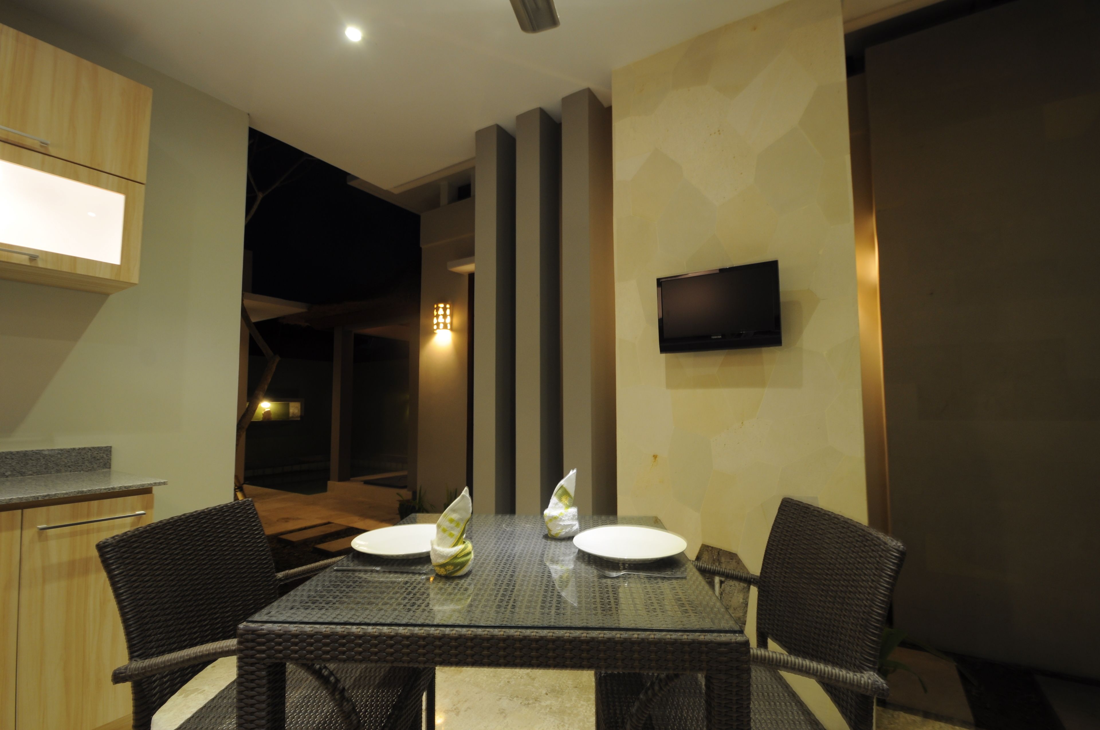 Un coin repas avec une table dressée pour deux personnes, un téléviseur à écran plat fixé au mur et un espace cuisine doté de meubles en bois et d’un plan de travail.