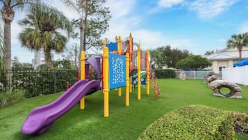 Children can play on the playground's slide and structures surrounded by lush grass and palm trees.