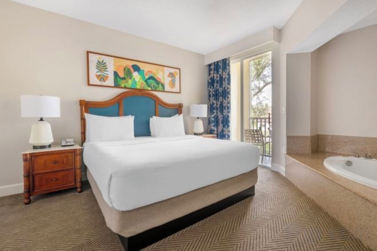 A hotel room with a large bed, two bedside tables with lamps, a painting on the wall, a balcony with a view, and a bathtub.