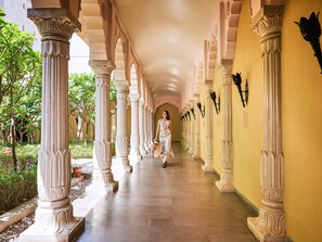 Un pasillo con columnas, una mujer caminando y una pared amarilla.