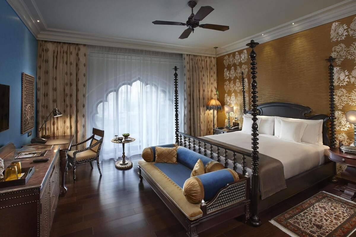 A four-poster bed with a canopy, a sitting area with a sofa and armchairs, a desk with a lamp, and a large window with curtains.