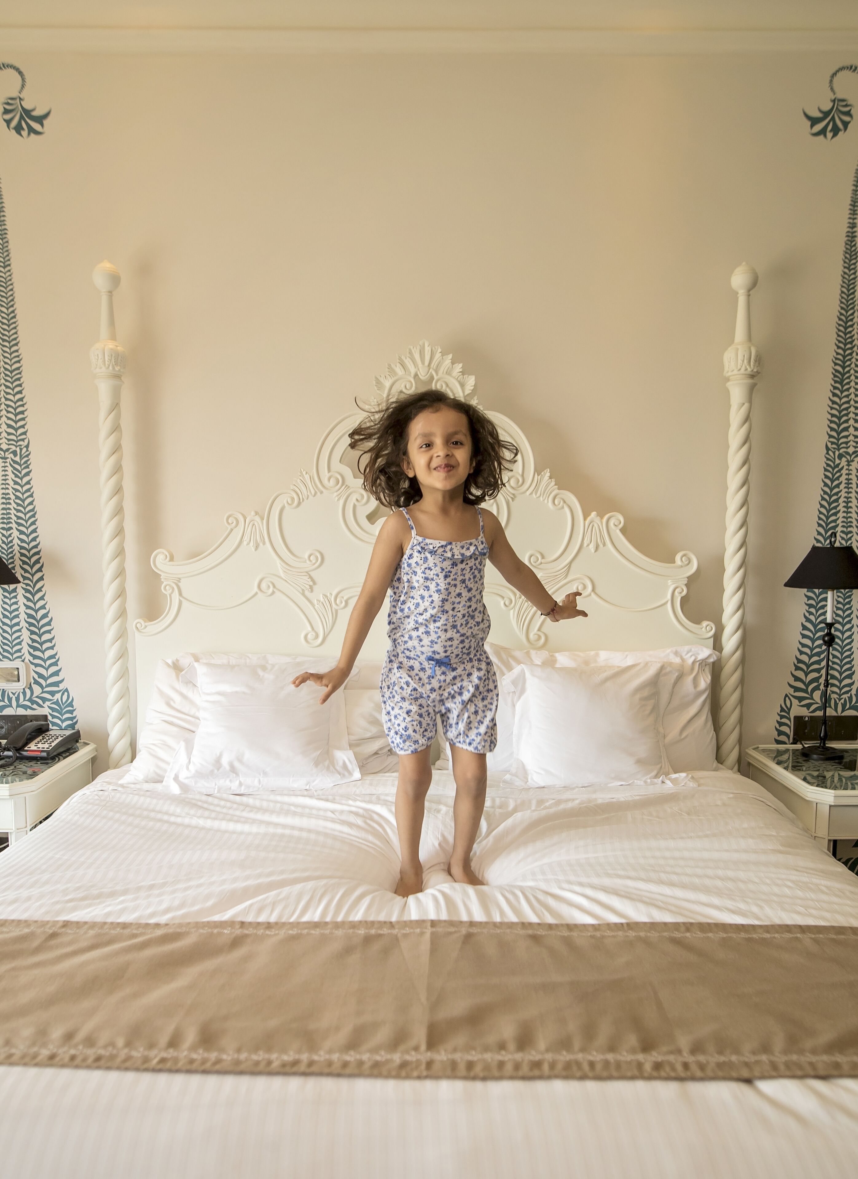 Un niño saltando sobre una cama con cabecera blanca y motivos de hojas decorativas en las paredes.