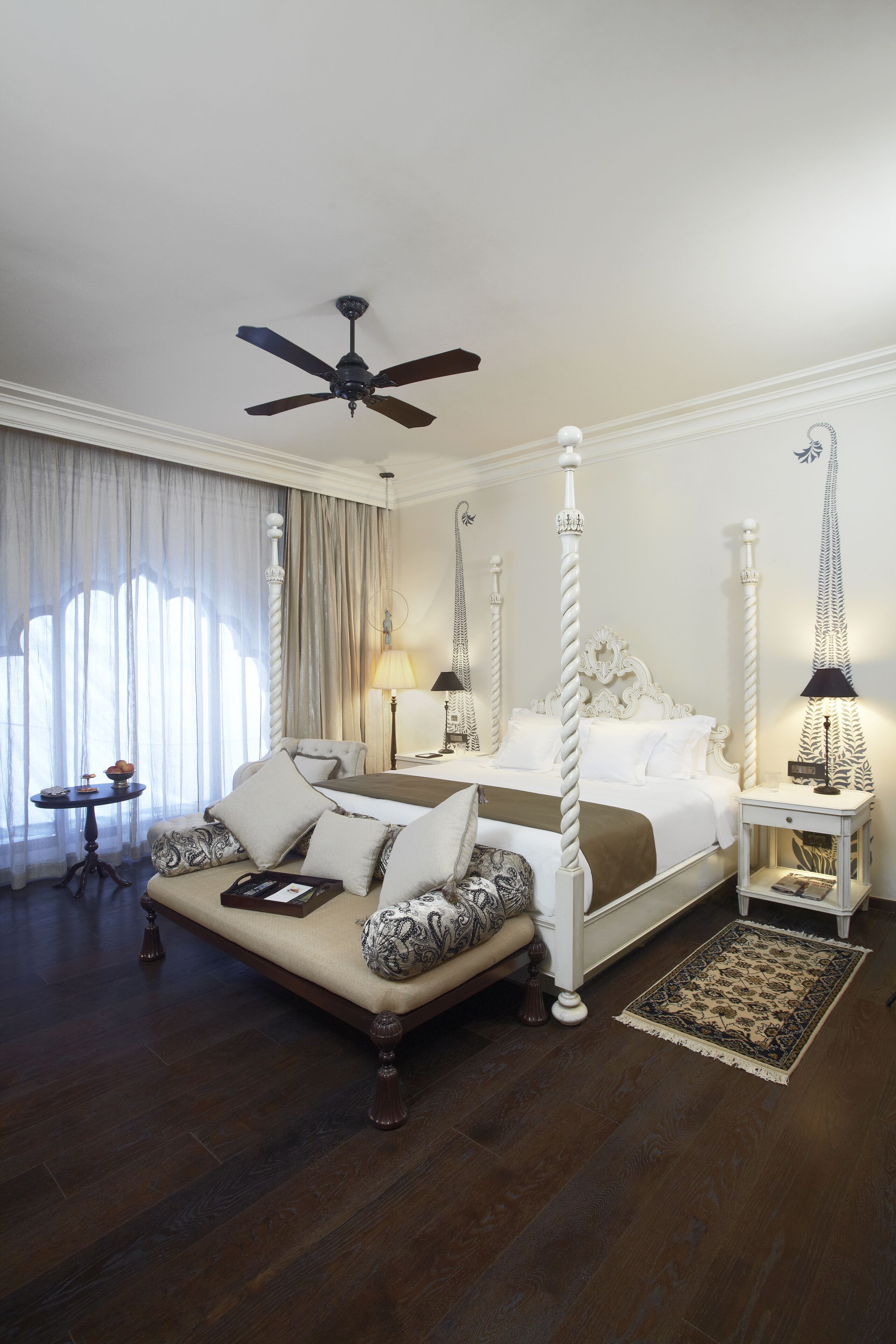 A four-poster bed with a canopy, a sitting area with a cushioned bench, a bedside table with a lamp, and a large window with curtains.