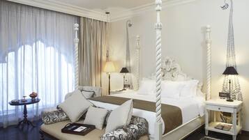 A four-poster bed with a canopy, a sitting area with a cushioned bench, a bedside table with a lamp, and a large window with curtains.