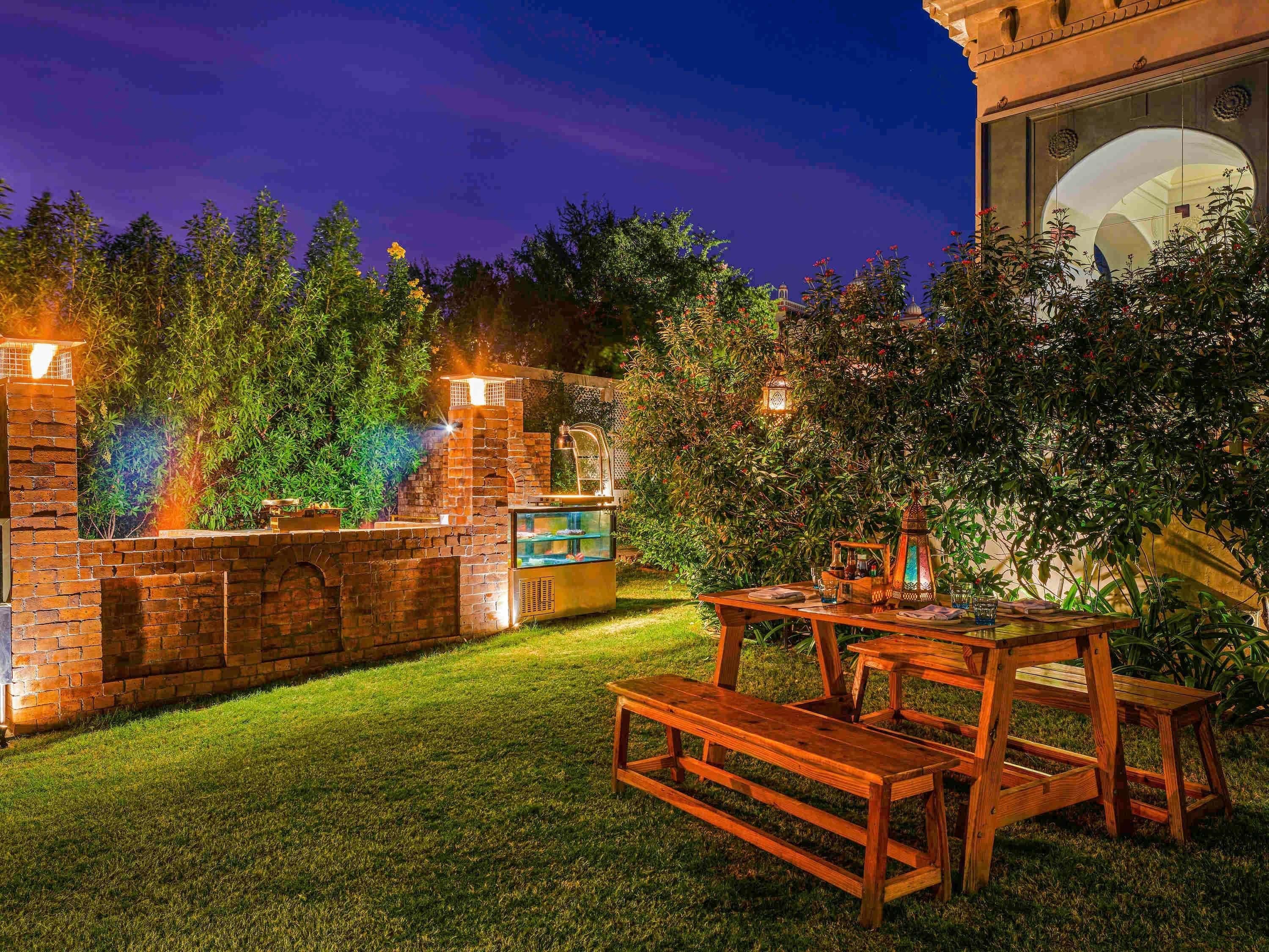 An outdoor patio with a brick barbecue area, a wooden table with benches, and a decorative lantern.