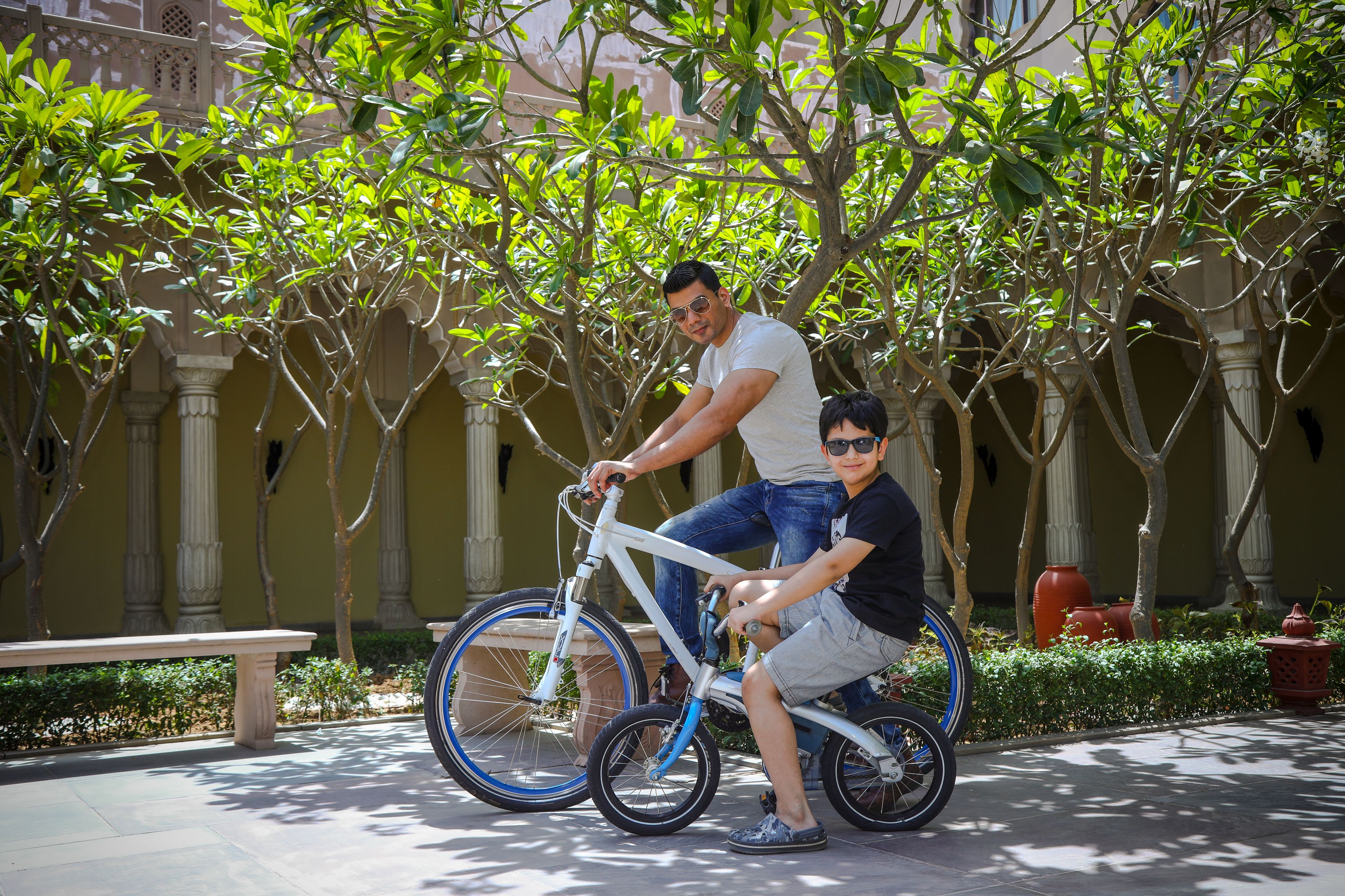 Un hombre y un niño en una bicicleta blanca en un patio con columnas y follaje.