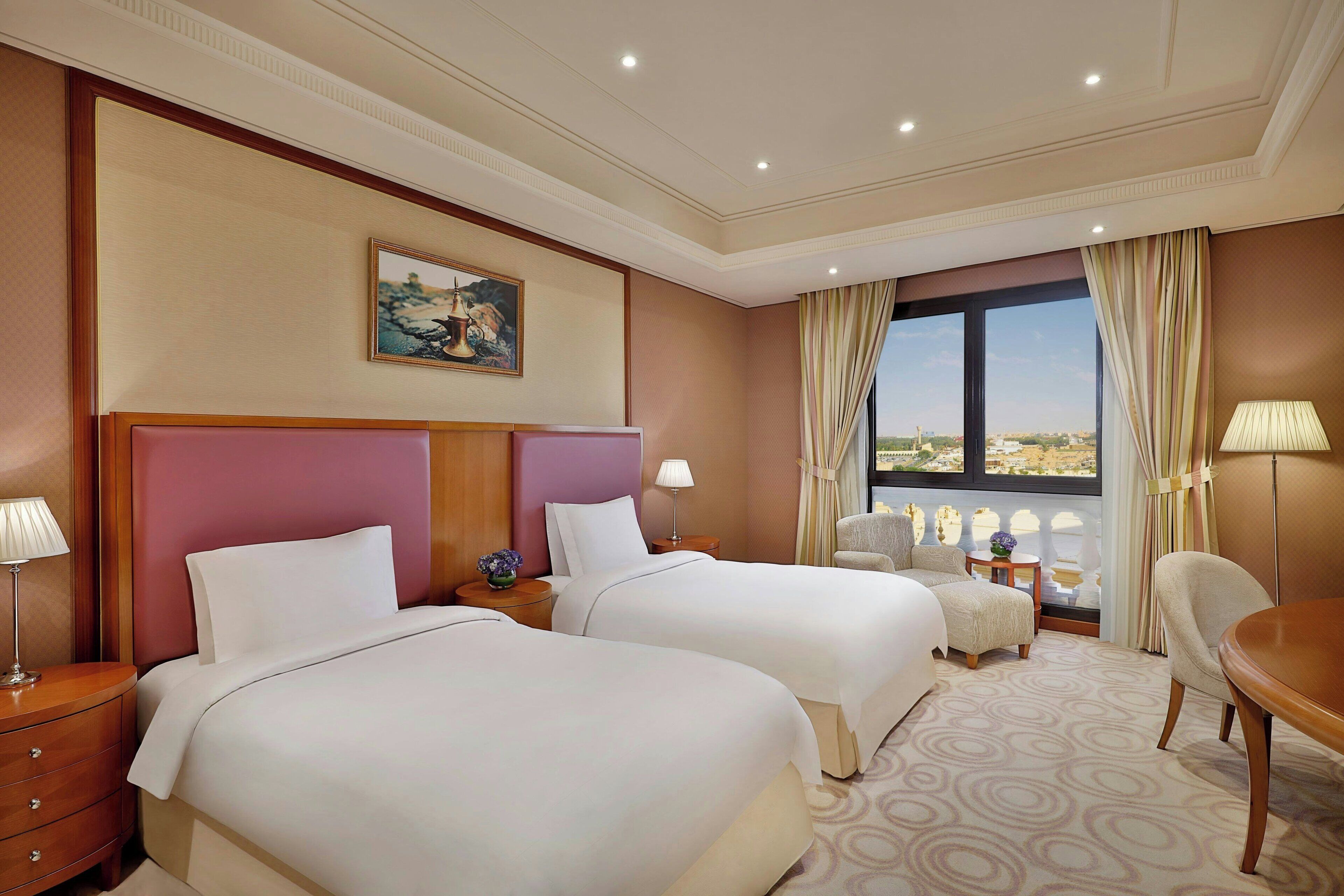 A hotel room with two beds, a wooden headboard, a painting, a view through the window, and a dining table.