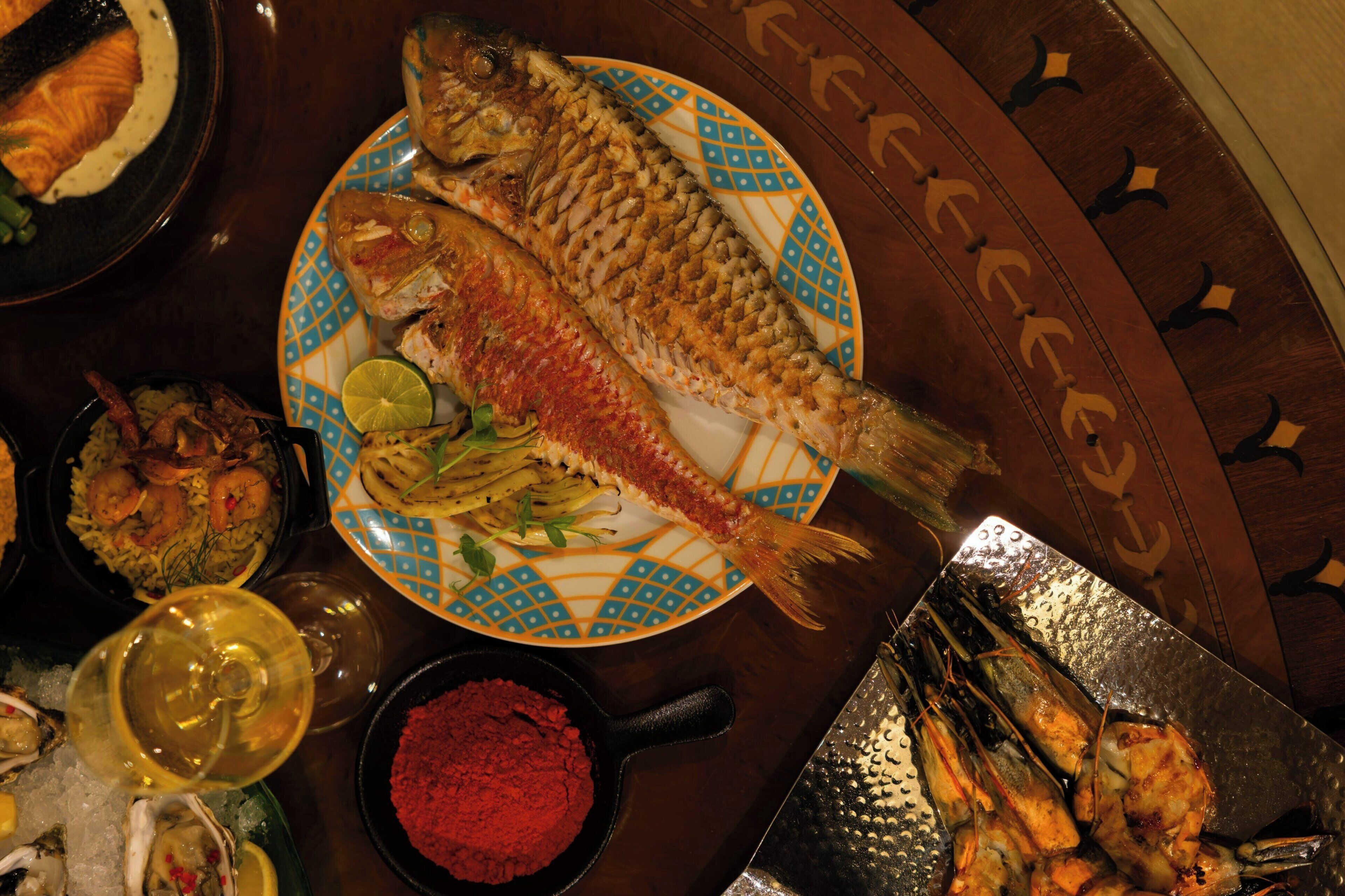 A dining table with a variety of seafood dishes, including a whole fish on a decorative plate, shrimp, and other seafood.