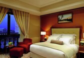 A hotel room with a large bed, a red armchair, a small table with a fruit bowl, a nightstand with a lamp, and a painting on the wall.