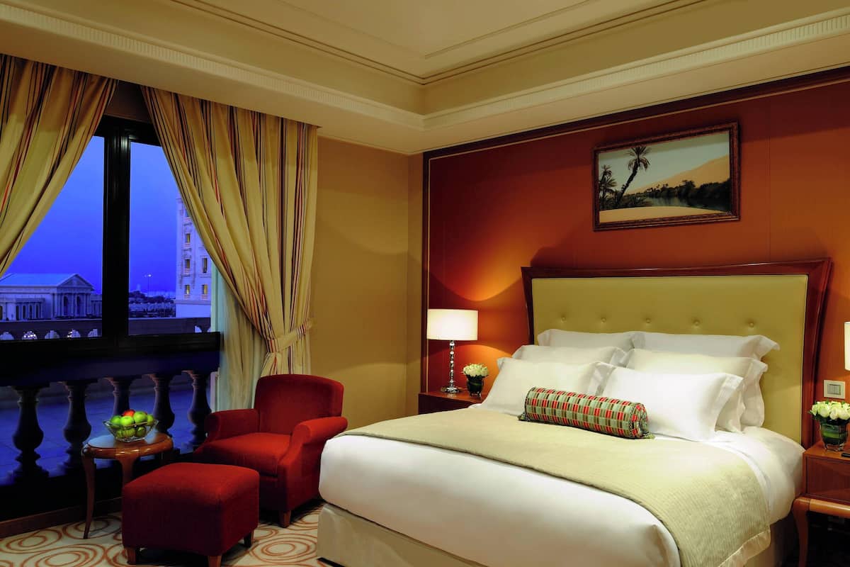 Quarto de hotel com uma cama grande, uma poltrona vermelha, uma mesinha com uma fruteira, uma mesa de cabeceira com abajur e um quadro na parede.